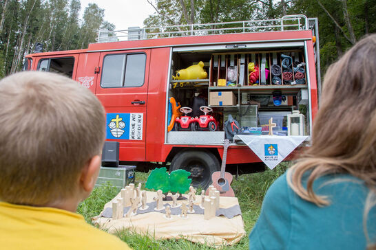 Perspektive über die Schultern von Kindern zu einem Feuerwehrauto, Türen sind geöffnet, im Innern sind BobbyCars. Vor dem Feuerauto ist eine Szene mit Figuren aus Holz aufgebaut.